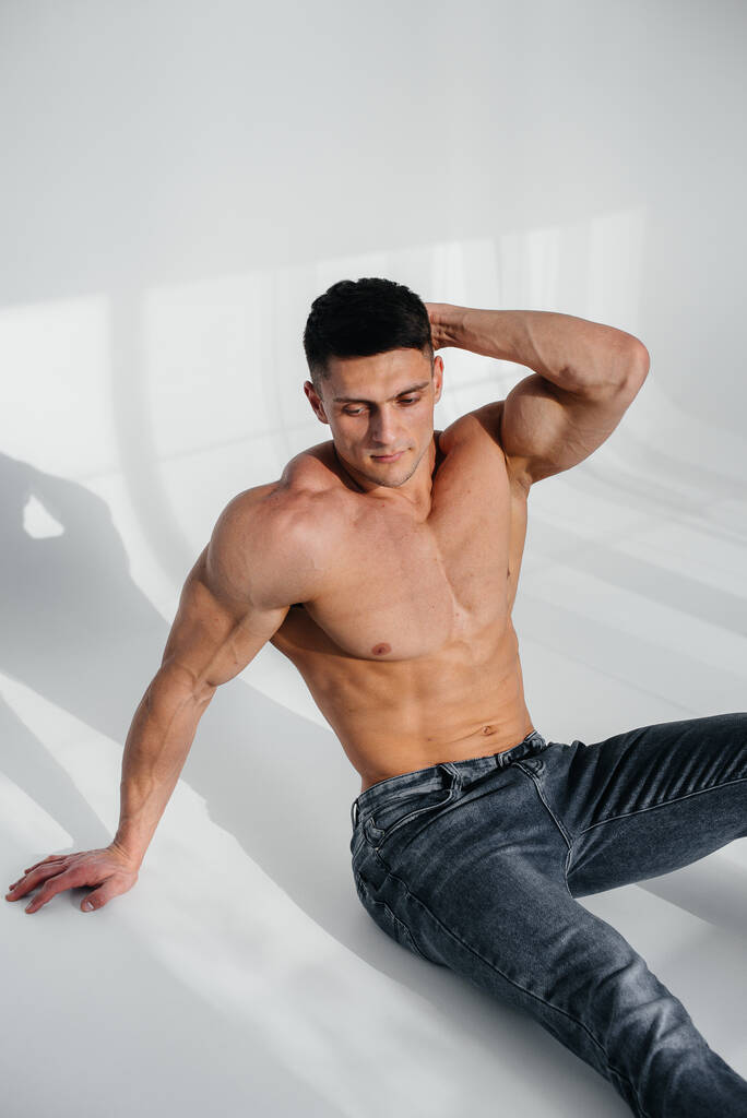 Um jovem atleta sexy com abdominais perfeitos senta-se no chão em topless estúdio em jeans no fundo. Estilo de vida saudável, nutrição adequada, programas de treinamento e nutrição para perda de peso. - Foto, Imagem