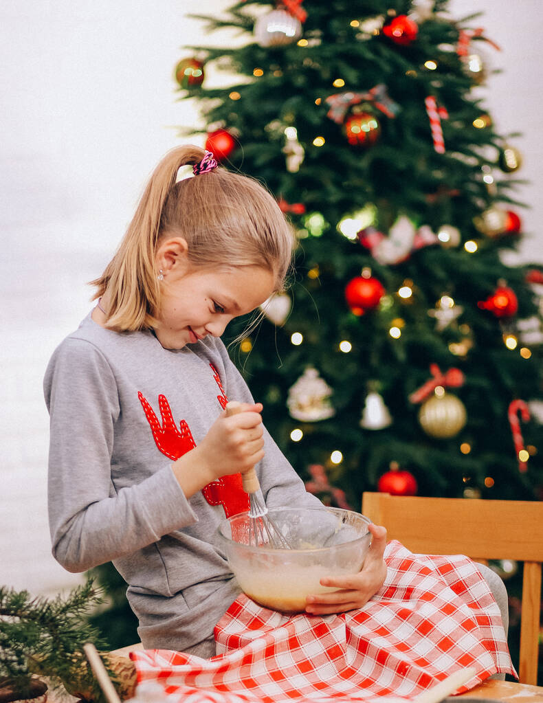 κορίτσι μαστιγώνει ένα ασπράδι αυγού με ένα σύρμα για να κάνει μπισκότα στο φόντο μιας χριστουγεννιάτικης tre - Φωτογραφία, εικόνα
