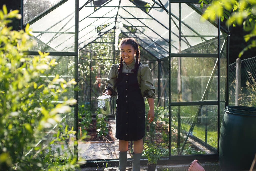 Happy small girl gardening in greenhouse outdoors in backyard, looking at camera. - Photo, Image