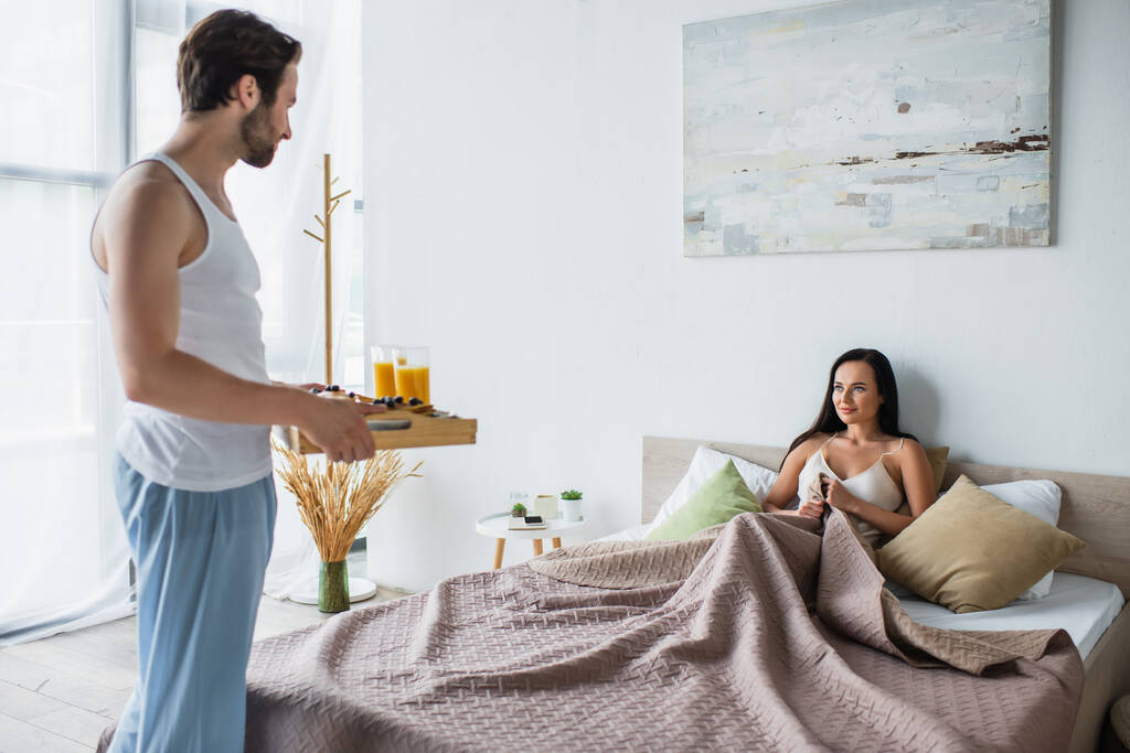 Bärtiger Mann hält Frühstückstablett neben lächelnder Freundin im Bett - Foto, Bild