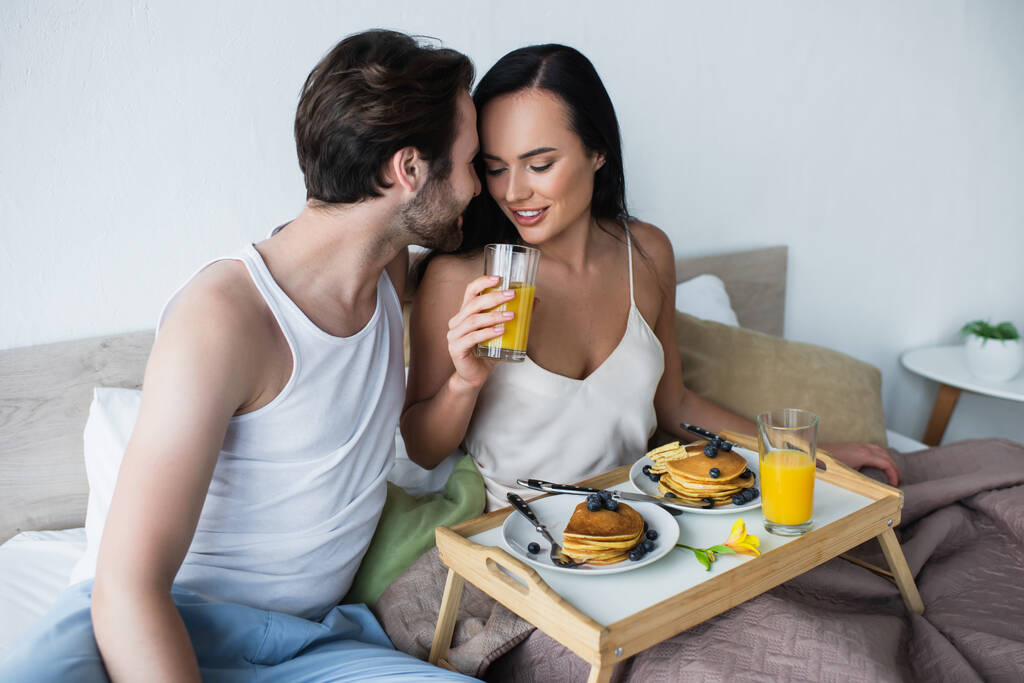 бородатый мужчина смотрит на счастливую девушку со стаканом апельсинового сока в постели - Фото, изображение