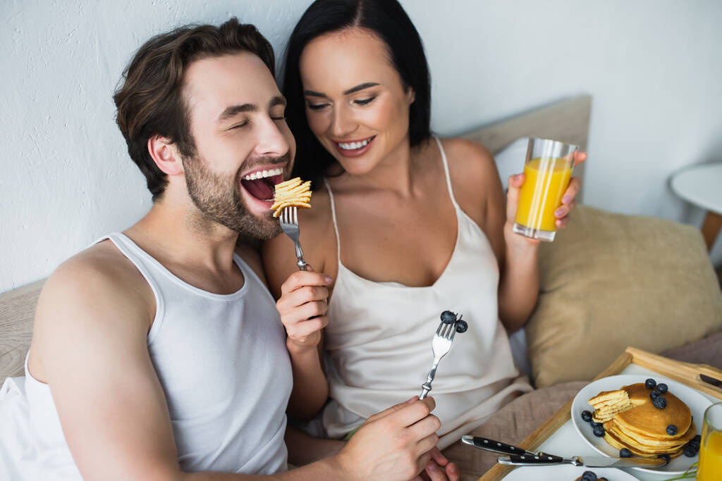 femme souriante nourrissant petit ami avec des crêpes savoureuses et tenant un verre de jus d'orange - Photo, image