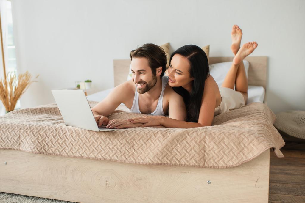 ベッドに横になりながらノートパソコンで映画を見る幸せなカップル  - 写真・画像