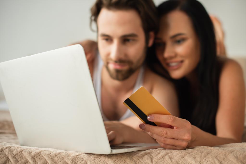 pareja borrosa celebración de la tarjeta de crédito cerca del ordenador portátil, mientras que las compras en línea en el dormitorio - Foto, Imagen