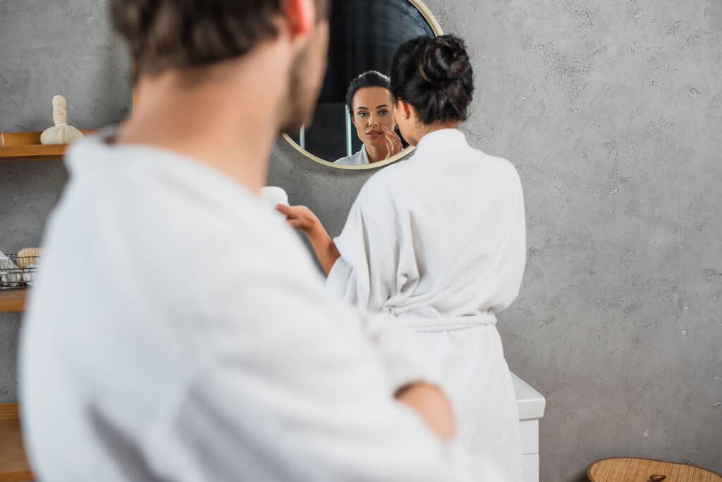 женщина наносит крем для лица и смотрит на зеркало рядом с размытым парнем на переднем плане  - Фото, изображение