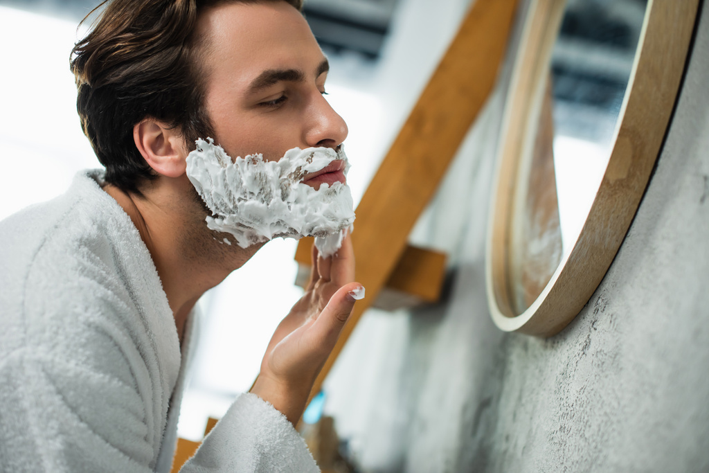 άνθρωπος εφαρμογή κρέμα ξυρίσματος στο πρόσωπο κοντά στον καθρέφτη - Φωτογραφία, εικόνα
