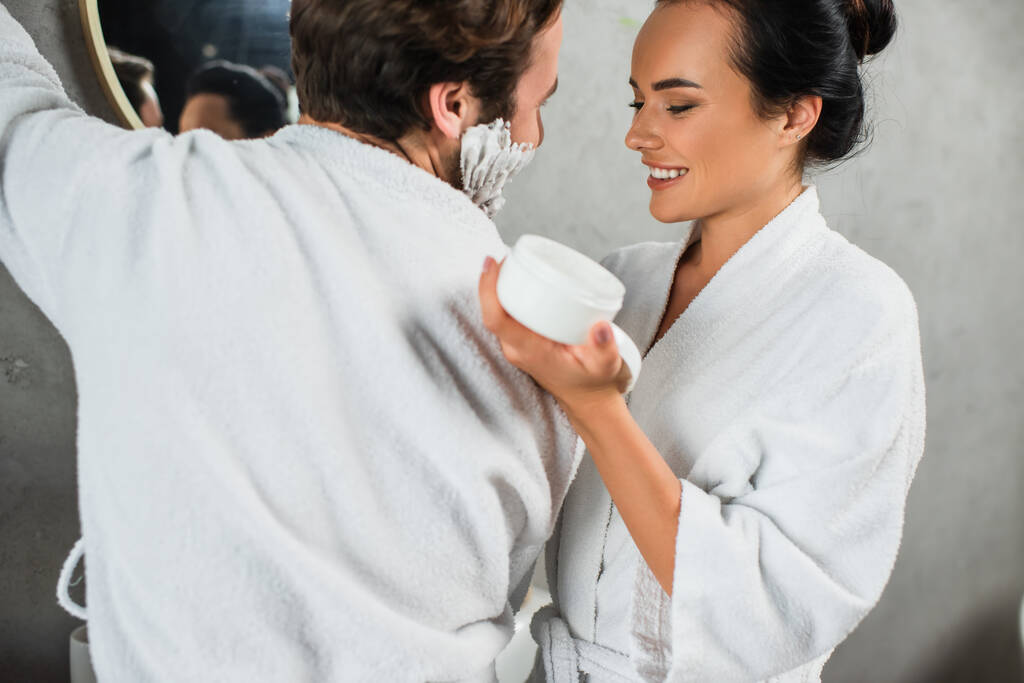 fröhliche Frau hält Behälter mit Gesichtscreme in der Nähe glücklicher Mann mit Rasierschaum im Gesicht - Foto, Bild