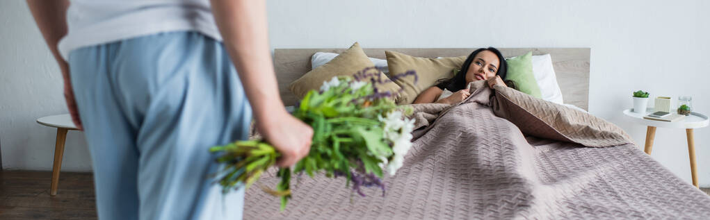 мужчина держит букет цветов рядом с подругой лежит на кровати, баннер - Фото, изображение