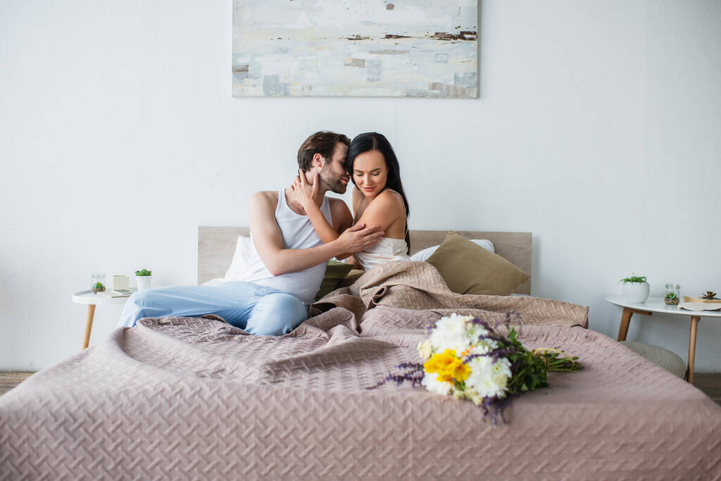 Blumenstrauß im Bett neben zufriedenem Paar, das sich auf dem Bett umarmt - Foto, Bild