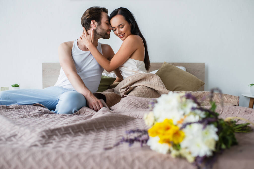 ramo borroso de flores cerca de feliz pareja abrazándose en la cama - Foto, Imagen