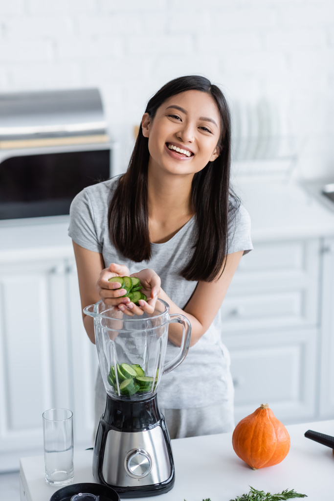 radostná asijská žena přidává nakrájenou okurku do mixéru a připravuje snídani v kuchyni - Fotografie, Obrázek