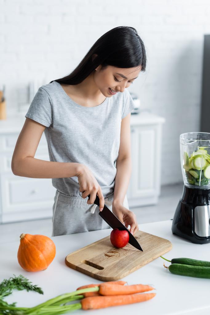 positivo asiático mujer corte manzana cerca eléctrico licuadora y verduras en cocina mesa - Foto, imagen