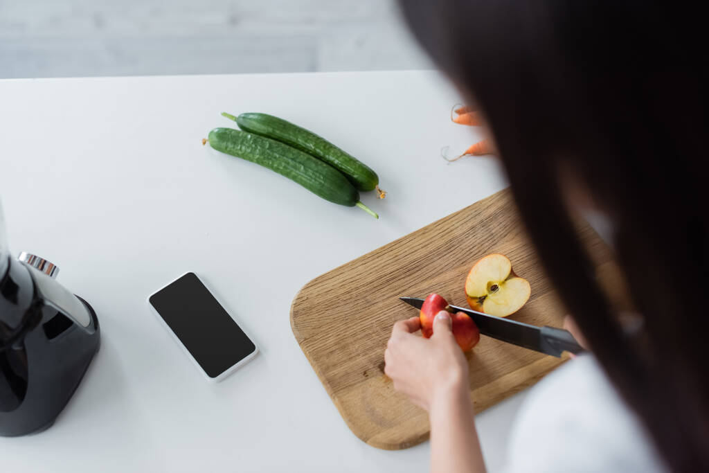 bovenaanzicht van wazige vrouw snijden appel in de buurt van smartphone met blanco scherm en verse komkommers - Foto, afbeelding