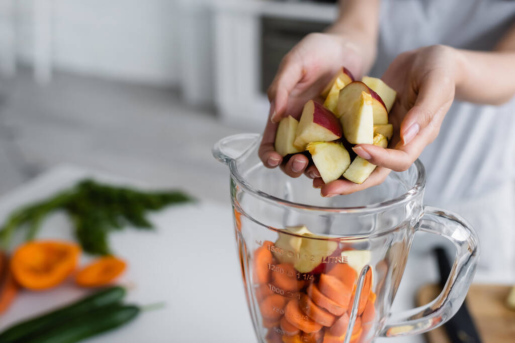 visão cortada de mulher adicionando maçãs em jarro de liquidificador com cenouras cortadas - Foto, Imagem