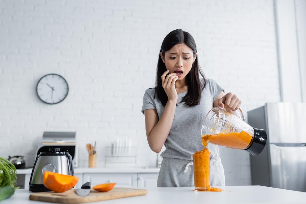 worried asian woman pouring smoothie into overflowing glass near cut pumpkin on chopping board - Photo, Image