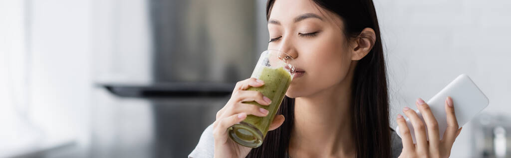 pleased asian woman with smartphone drinking tasty smoothie with closed eyes, banner - Photo, Image