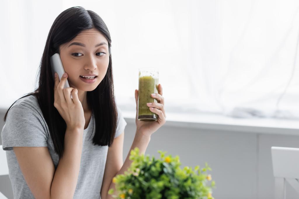 nuori aasialainen nainen soittaa matkapuhelimeen pitäen lasin smoothie lähellä hämärtynyt kasvi - Valokuva, kuva