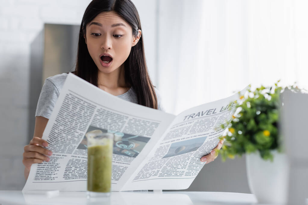 изумленная азиатка читает газету "Жизнь во время путешествий" возле размытого стекла с смузи - Фото, изображение