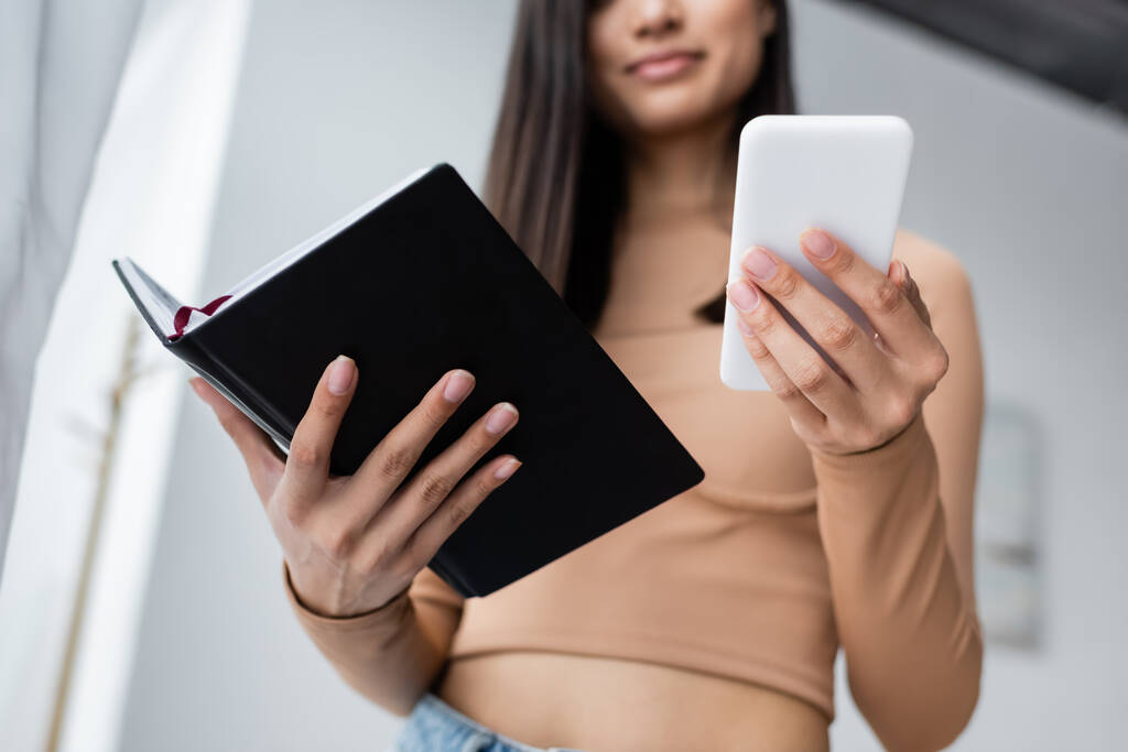 cropped view of blurred woman holding smartphone and notebook while working at home - Photo, Image