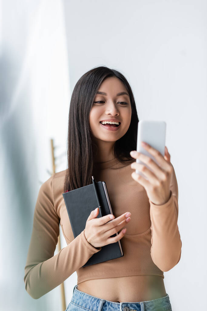 веселая азиатка, держащая ноутбук и указывая пальцем во время видеозвонка на смартфон - Фото, изображение