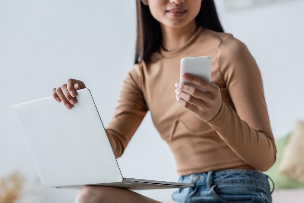 vista cortada de mulher jovem usando smartphone perto de laptop em casa - Foto, Imagem