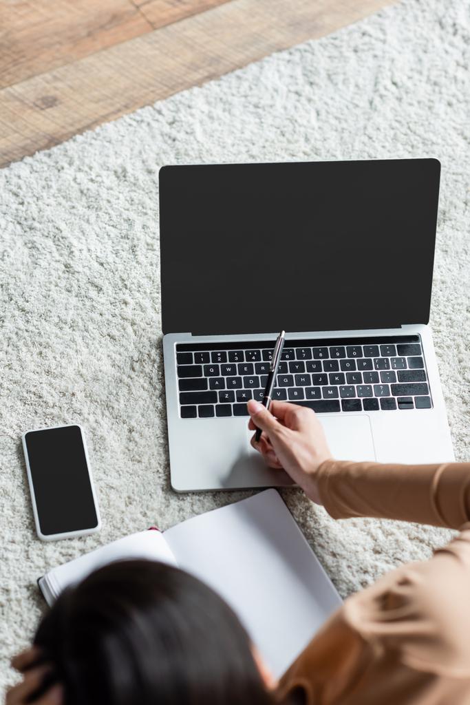 widok z góry rozmyty kobieta wskazując piórem na laptopa w pobliżu smartfona z pustym ekranem na podłodze - Zdjęcie, obraz