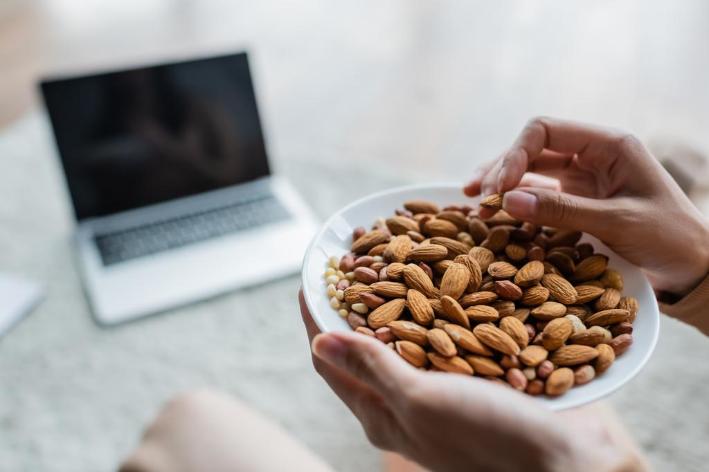 partial view of woman with bowl of almonds near blurred laptop with blank screen - Photo, Image