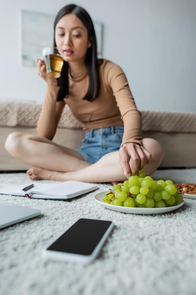 niewyraźne Azji kobieta biorąc winogron podczas picia herbaty na podłodze w pobliżu notebook i smartfon - Zdjęcie, obraz