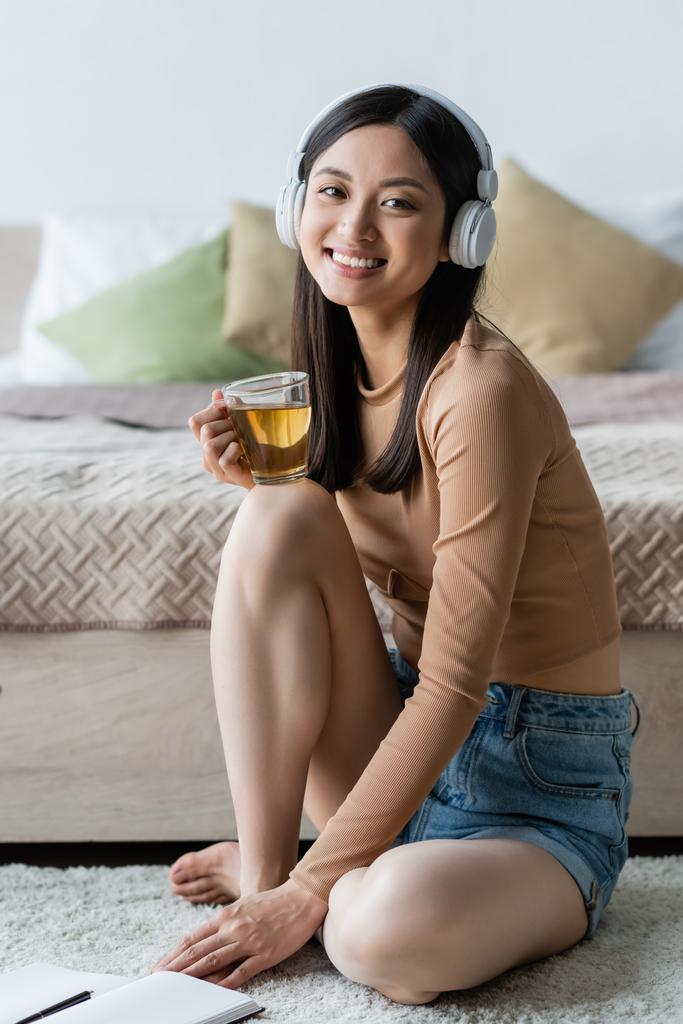 Ασιάτισσα γυναίκα με ακουστικά κρατώντας ένα φλιτζάνι τσάι και χαμογελώντας στην κάμερα στην κρεβατοκάμαρα - Φωτογραφία, εικόνα