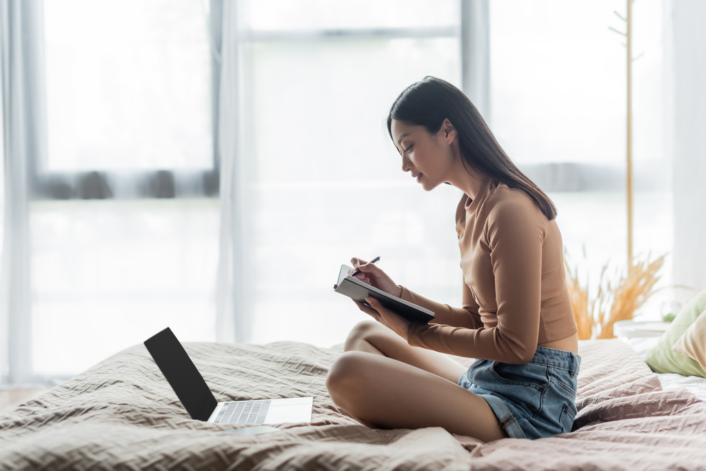 足を組んでベッドの上に座ってノートパソコンのそばにノートを書いているアジア人女性の姿です - 写真・画像