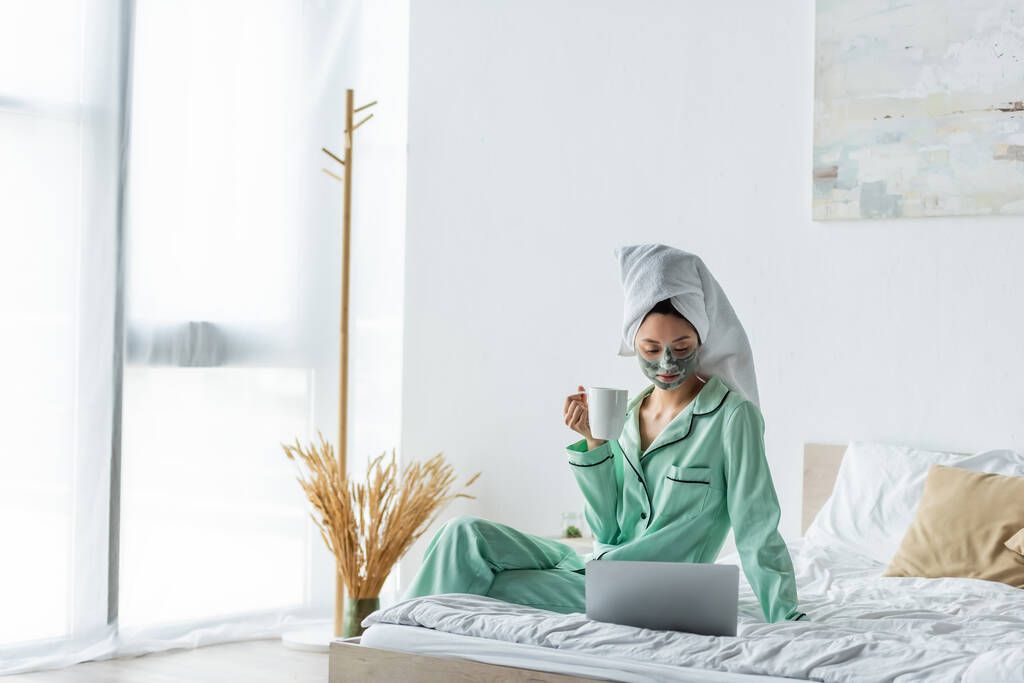 Aasialainen nainen savimaskissa ja pyjamassa istuu sängyllä teekupilla ja katselee kannettavaa tietokonetta - Valokuva, kuva
