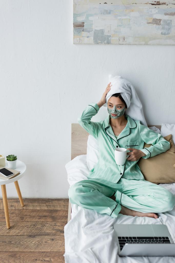 asiatique femme dans argile masque et pyjama toucher serviette sur la tête tandis que assis près de portable sur lit - Photo, image