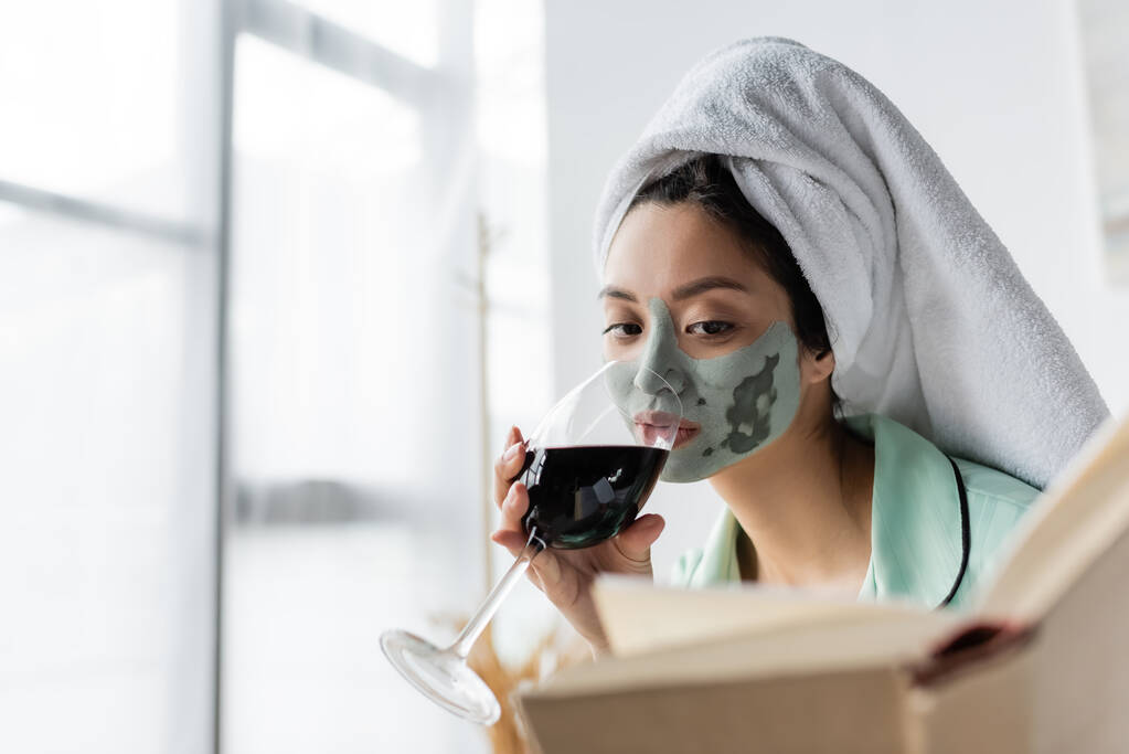 donna asiatica con maschera di argilla e asciugamano sulla testa bere vino rosso durante la lettura del libro a casa - Foto, immagini