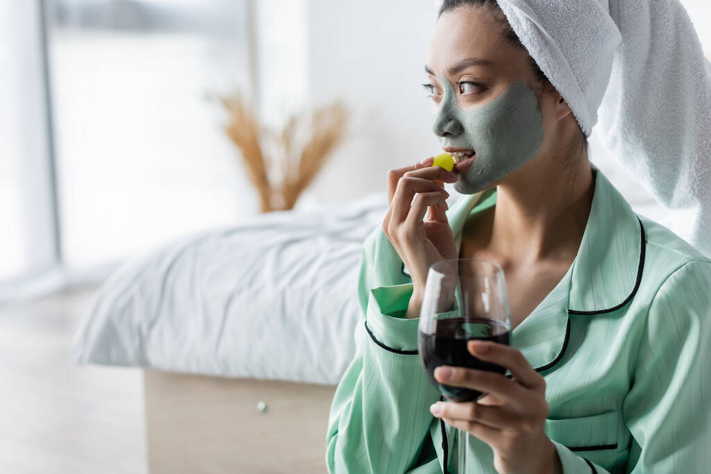 Ασιάτισσα με πήλινη μάσκα που τρώει σταφύλια κρατώντας ένα ποτήρι κόκκινο κρασί στην κρεβατοκάμαρα - Φωτογραφία, εικόνα