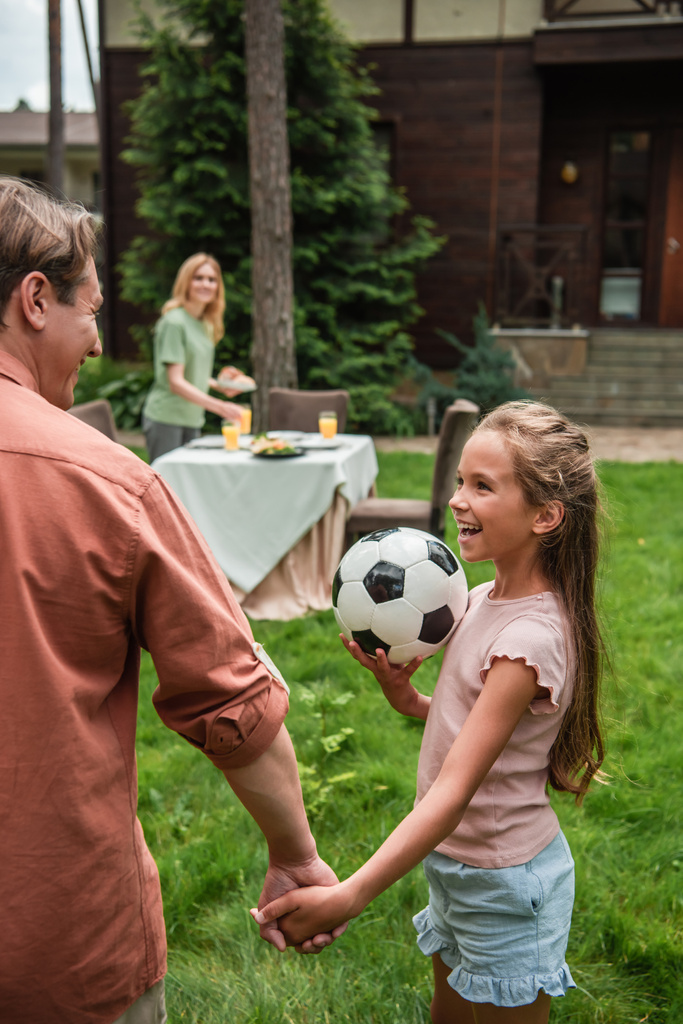 Sonriente niño sosteniendo pelota de fútbol y sosteniendo la mano del padre al aire libre  - Foto, imagen