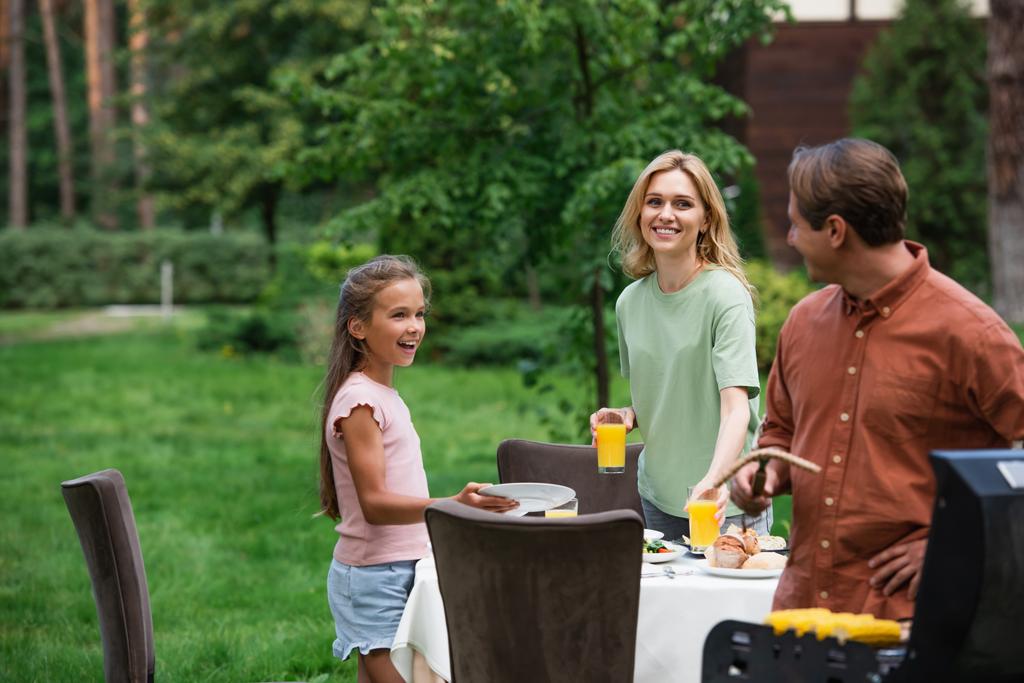 Smiling woman and girl looking at father grilling food outdoors  - Foto, afbeelding
