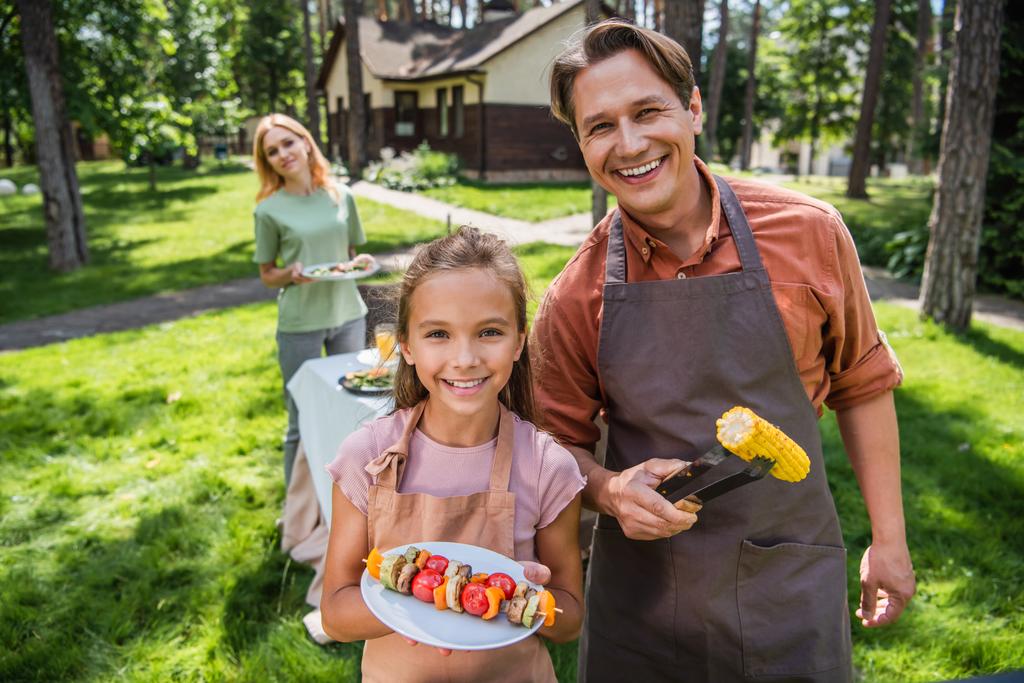 Mosolygó apa és gyerek kötényben kezében grillezett zöldségek a szabadban  - Fotó, kép