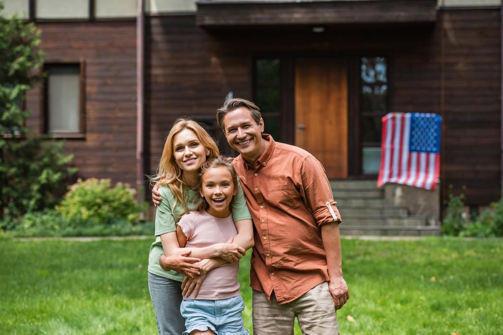 Sonriente familia abrazándose cerca borrosa bandera americana en casa al aire libre  - Foto, Imagen