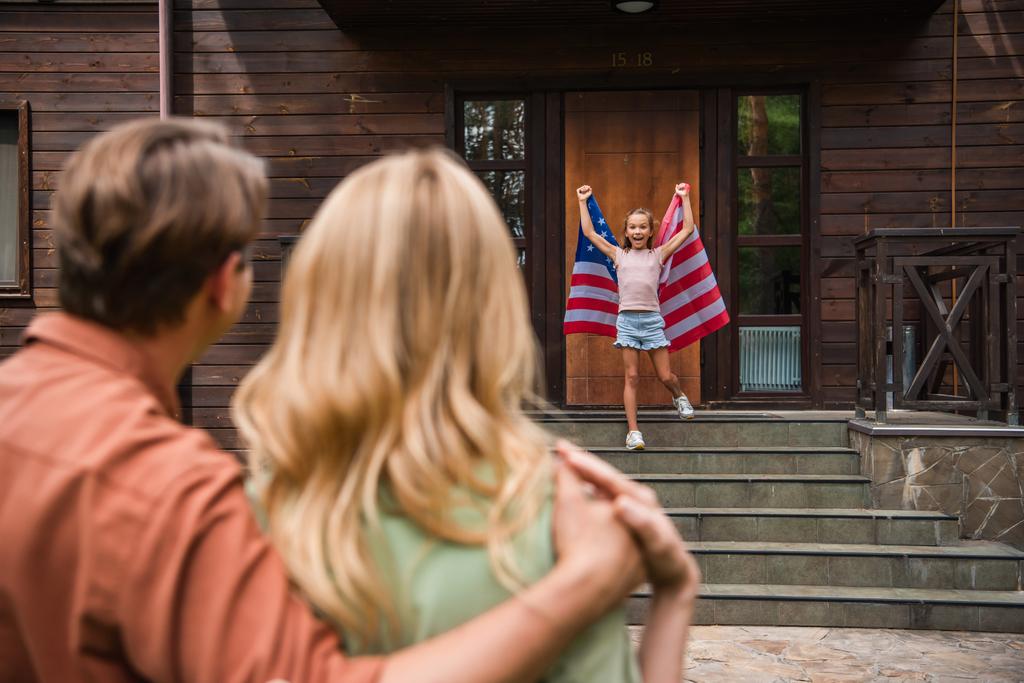 vzrušená dívka drží americkou vlajku v blízkosti rozmazané rodiče objímající venku  - Fotografie, Obrázek