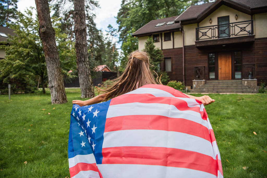 Rückansicht eines Mädchens mit amerikanischer Flagge beim Laufen auf dem Rasen  - Foto, Bild