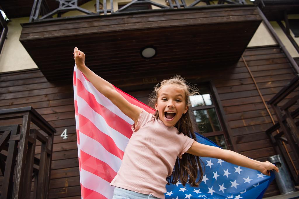 Garoto animado com bandeira americana olhando para a câmera perto da casa de férias  - Foto, Imagem