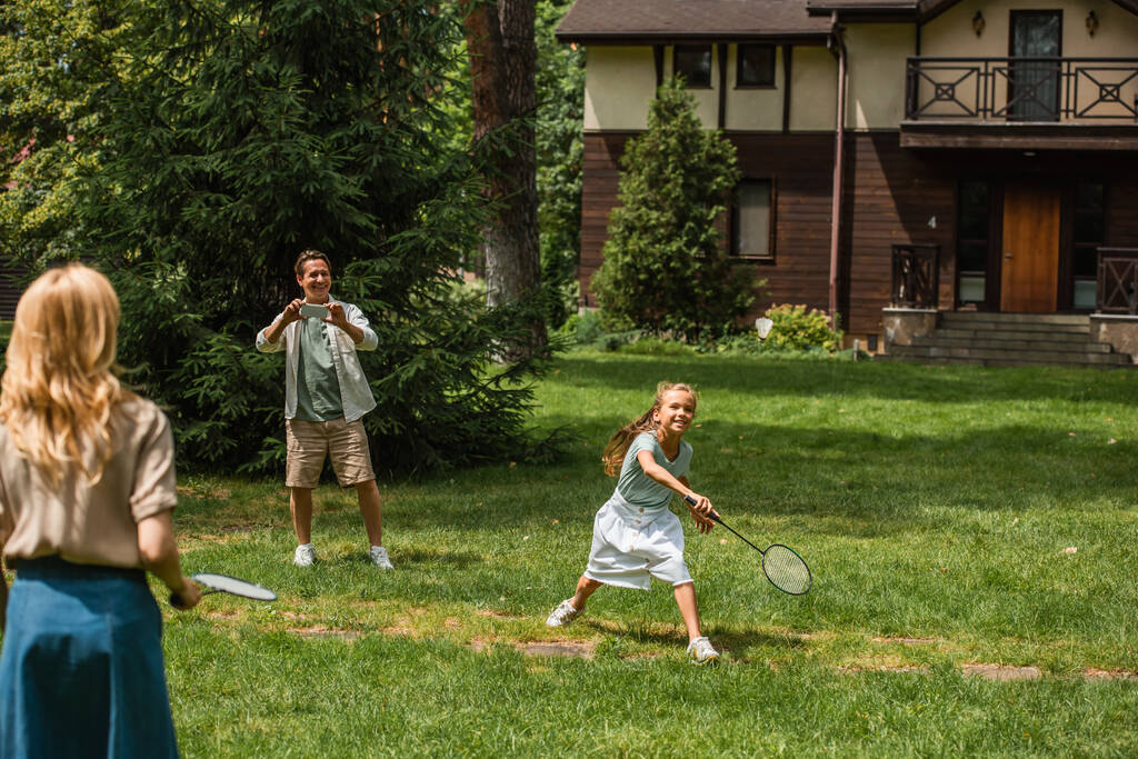 Kid i matka grają w badmintona w pobliżu ojca ze smartfonem na świeżym powietrzu  - Zdjęcie, obraz