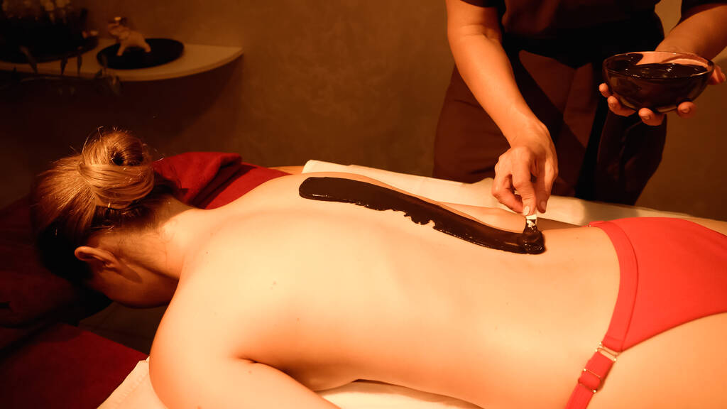 masażysta stosujący błoto ze szpatułką na ciele młodego klienta w salonie spa - Zdjęcie, obraz