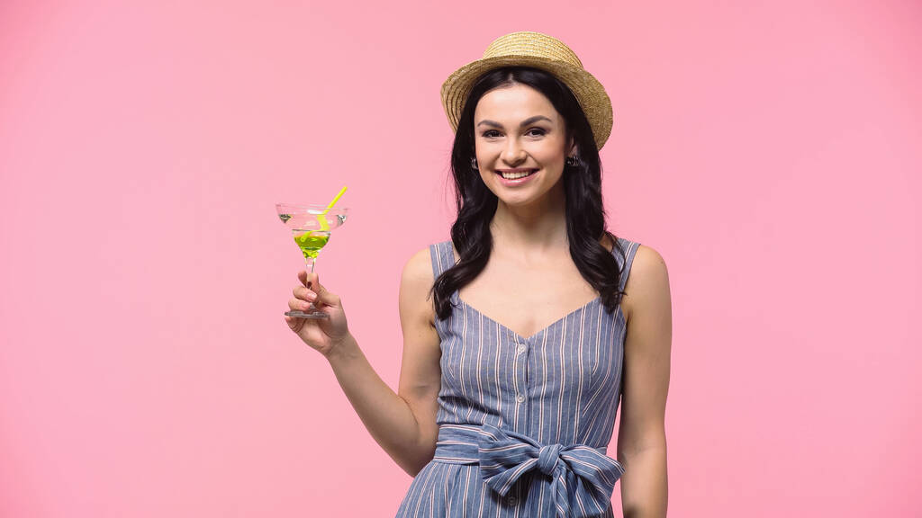 Positive Frau mit Sonnenhut hält Cocktail in der Hand und blickt vereinzelt in die Kamera auf rosa  - Foto, Bild