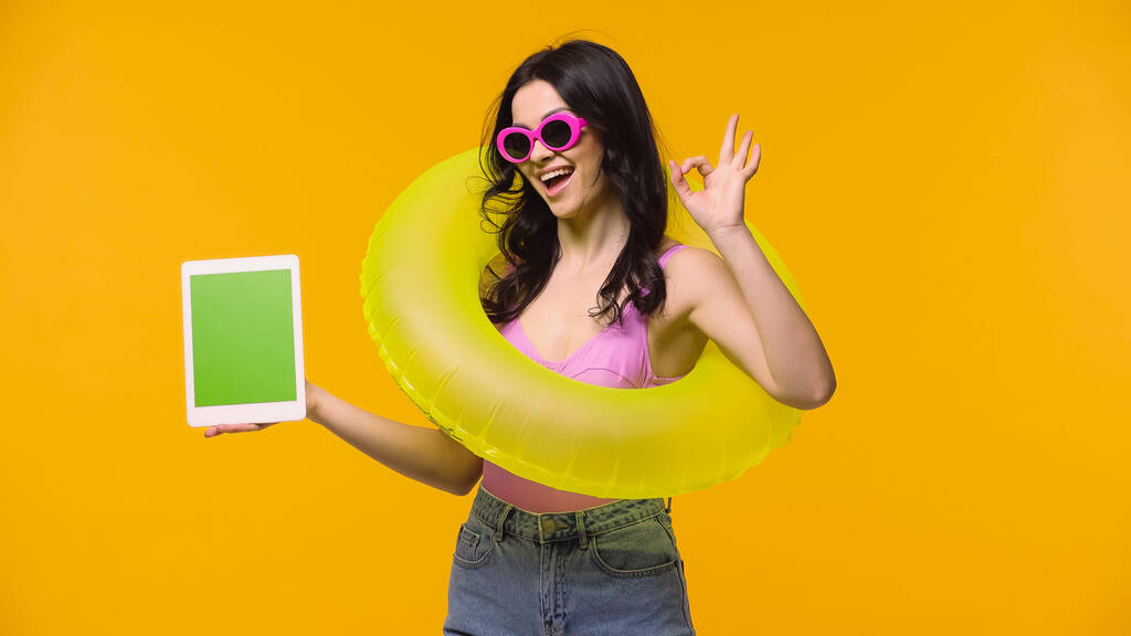 Nainen aurinkolasit ja uida rengas osoittaa ok pitäen digitaalinen tabletti vihreä näyttö eristetty keltainen  - Valokuva, kuva
