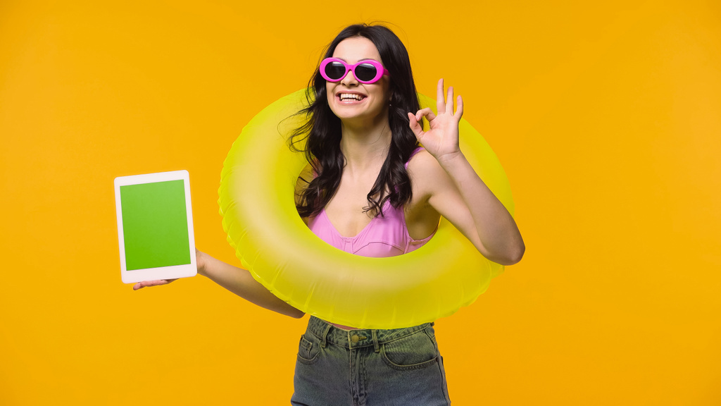 Uśmiechnięta kobieta w okularach przeciwsłonecznych i kostiumie kąpielowym trzymająca cyfrową tabletkę z zielonym ekranem i dobrze odizolowana na żółtym  - Zdjęcie, obraz