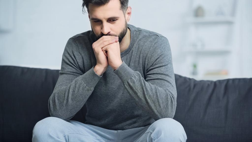 Besorgter Mann im grauen Pullover sitzt mit geballten Händen auf Couch - Foto, Bild