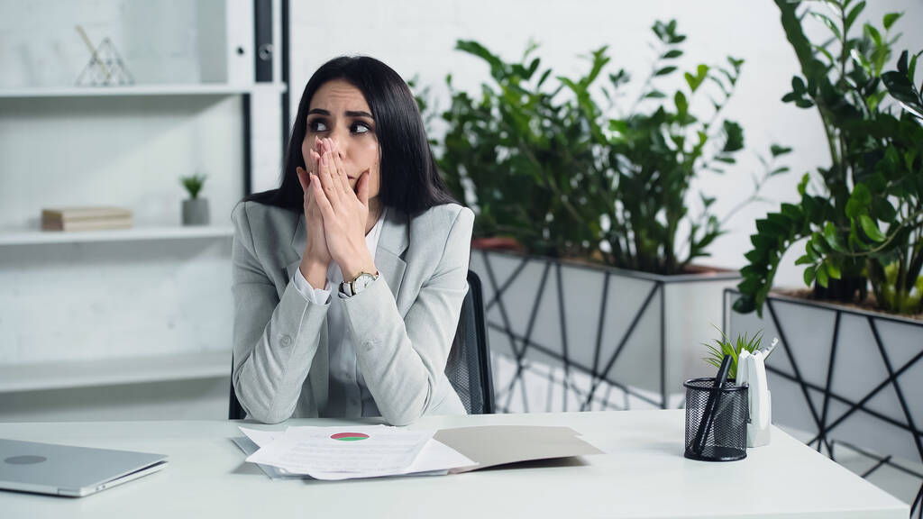 worried businesswoman looking away while sitting with praying hands near documents in office  - Photo, Image