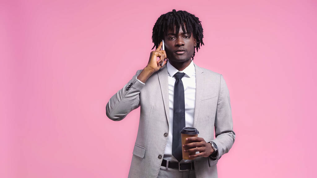 mladý africký americký podnikatel drží papírový šálek a mluví na mobilní telefon izolované na růžové - Fotografie, Obrázek