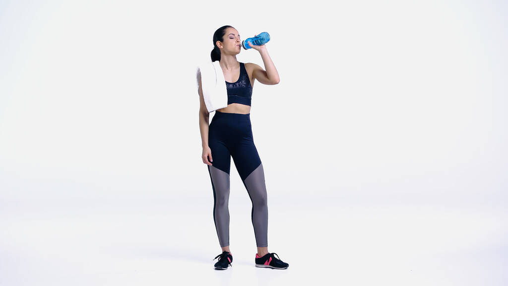 müde Sportlerin mit Handtuch trinkt Wasser aus Sportflasche auf weiß - Foto, Bild